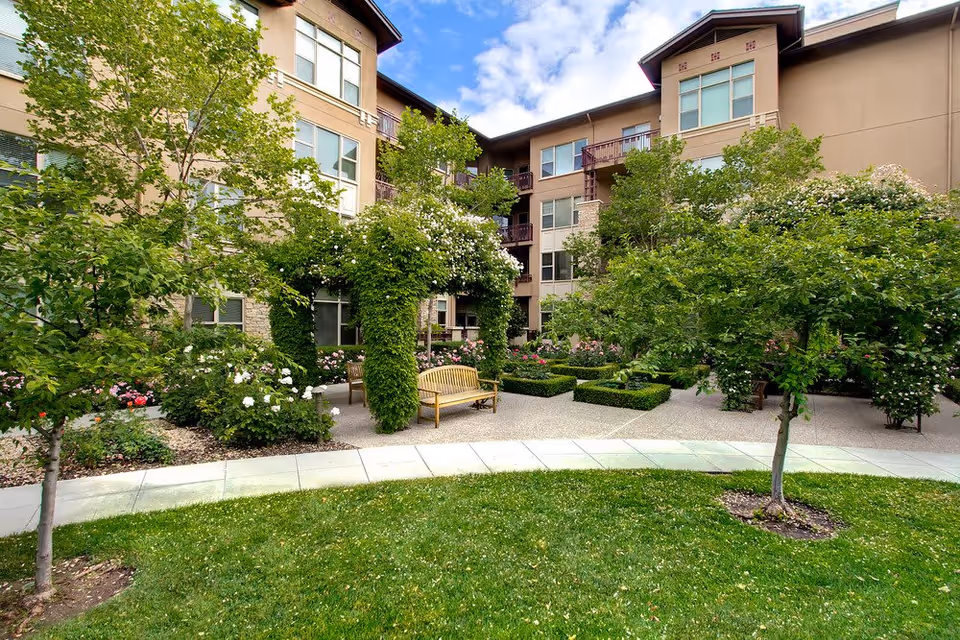 A landscaped courtyard with benches, flowering shrubs and trees in front of a multi-story residential building.