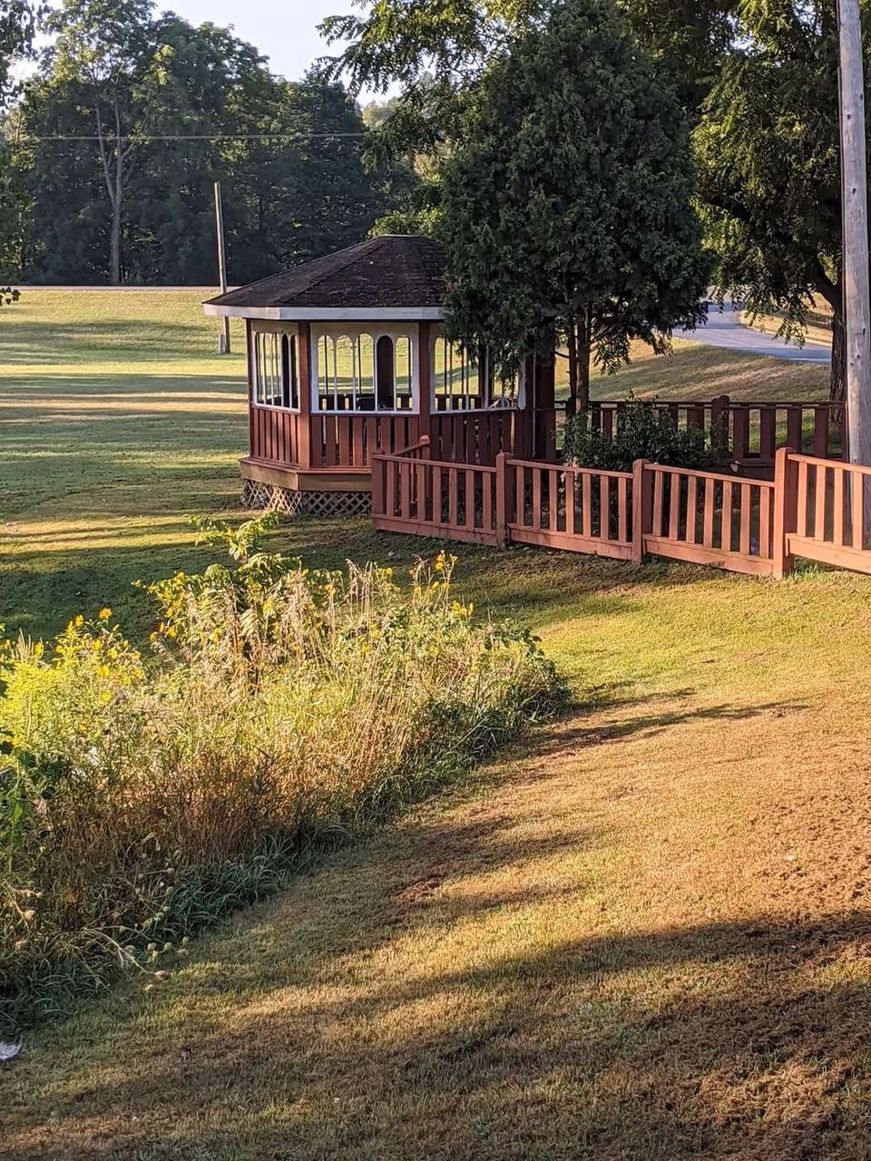 A small wooden gazebo with a shingled roof surrounded by a wooden fence on a grassy area with trees and bushes nearby. The scene is outdoors with sunlight casting shadows on the grass.