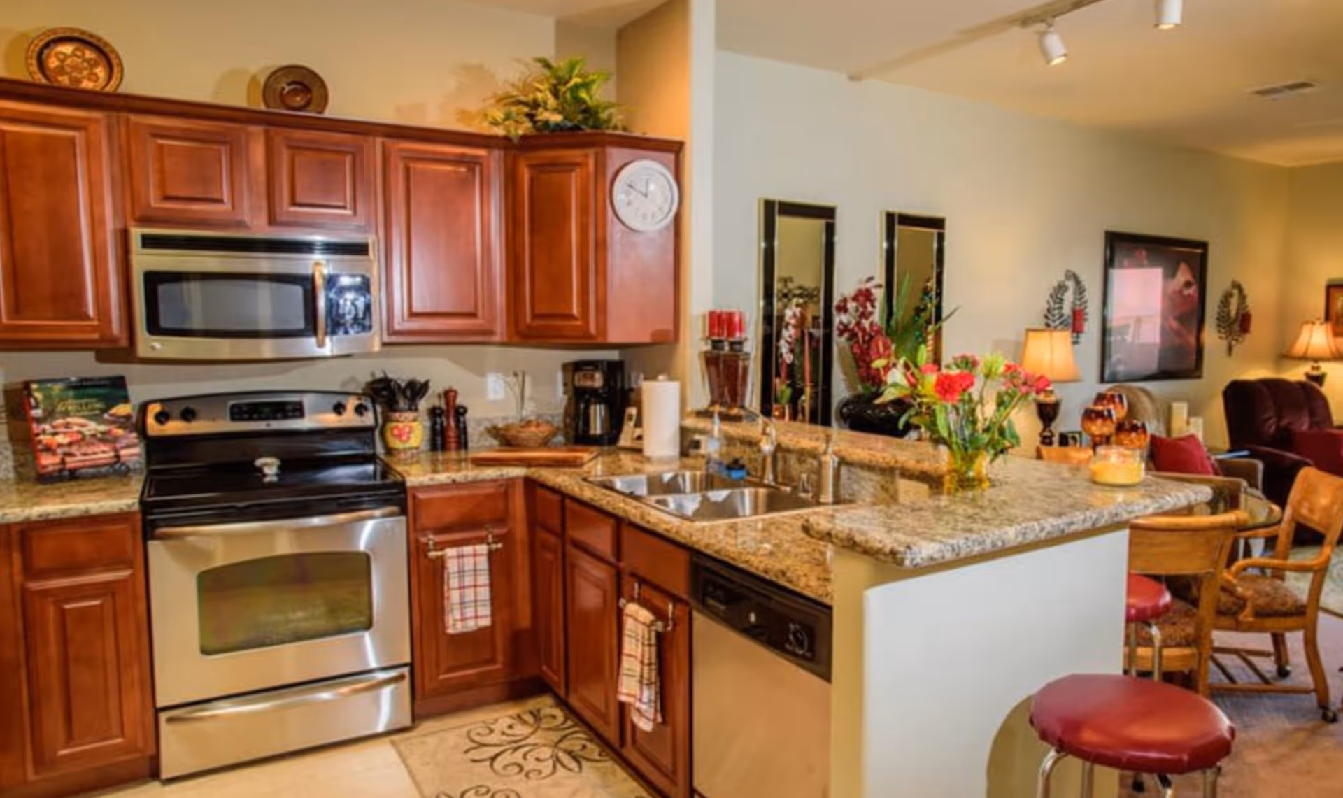 Open-plan kitchen with wooden cabinets, stainless steel stove and microwave, granite countertops and a breakfast bar with red stools opening to a living area.