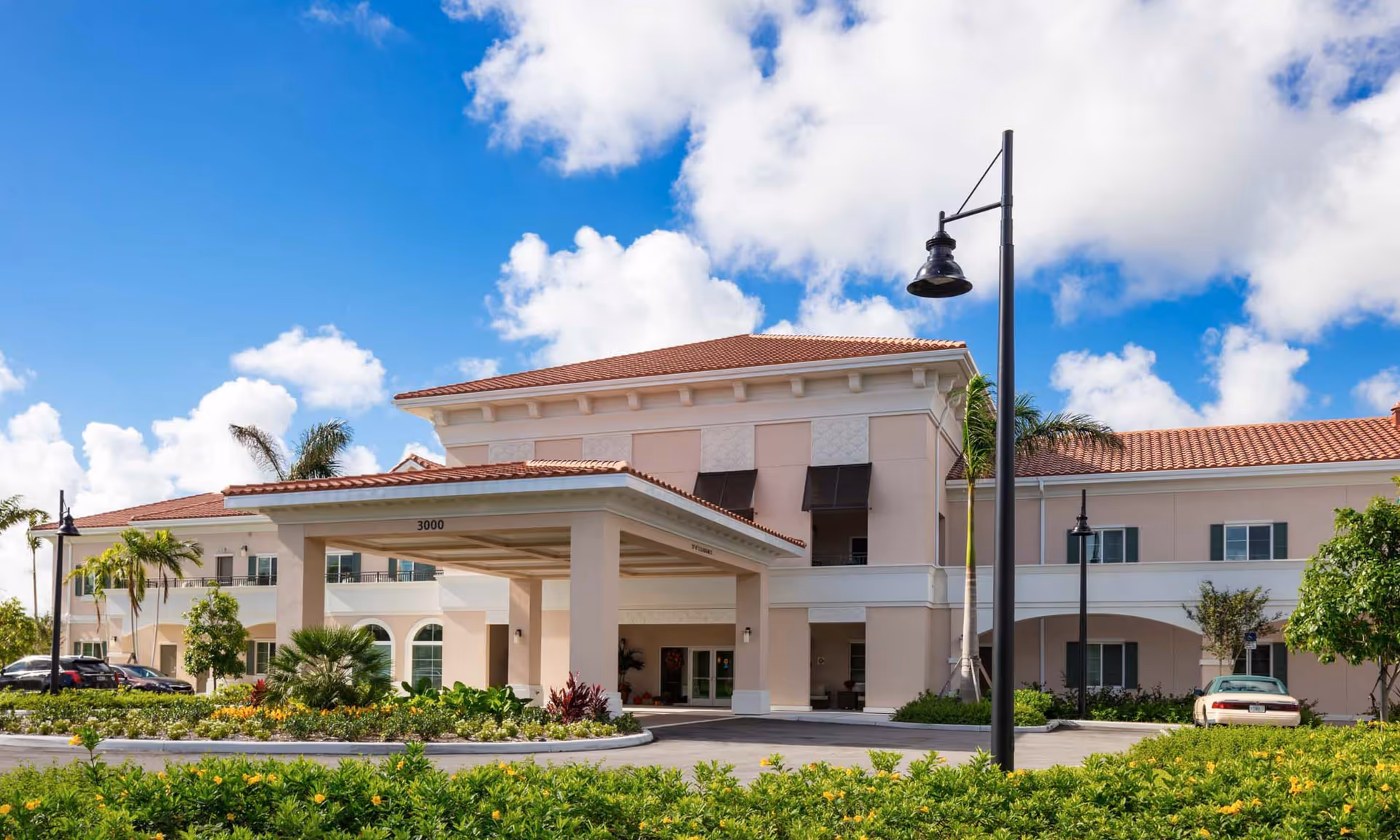 Exterior view of HarborChase of Palm Beach Gardens, a two-story building with a covered entrance, beige walls, red-tiled roof, surrounded by landscaped greenery and palm trees under a blue sky with scattered clouds.