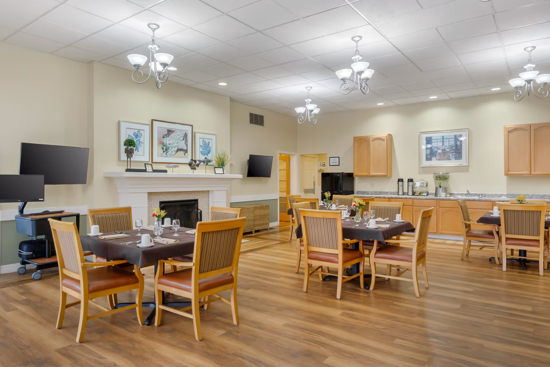 Bright dining room with several set tables and wooden chairs, a fireplace, wall-mounted TVs and a kitchenette area.