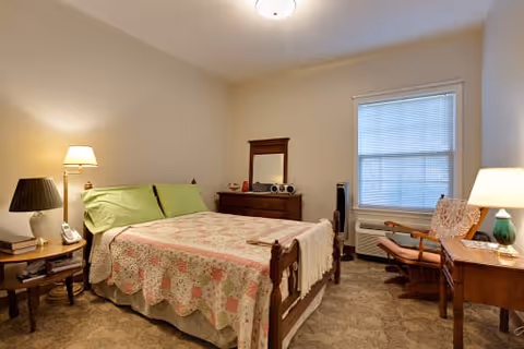 A cozy bedroom with a wooden bed frame covered by a floral quilt and green pillows. The room includes a wooden dresser with a mirror, a rocking chair near a window with blinds, a small wooden desk with a lamp, and a side table with a lamp and a phone. The carpet is patterned and the walls are painted light beige.
