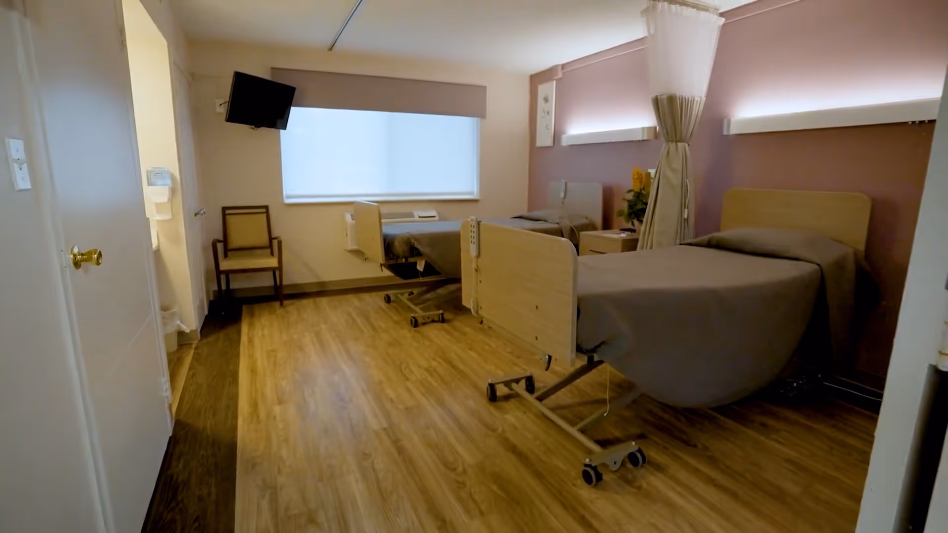 A healthcare facility room with two hospital beds separated by a privacy curtain. The room has wood-style flooring, a window with a blind, a wall-mounted TV, a chair, and soft lighting above the beds.