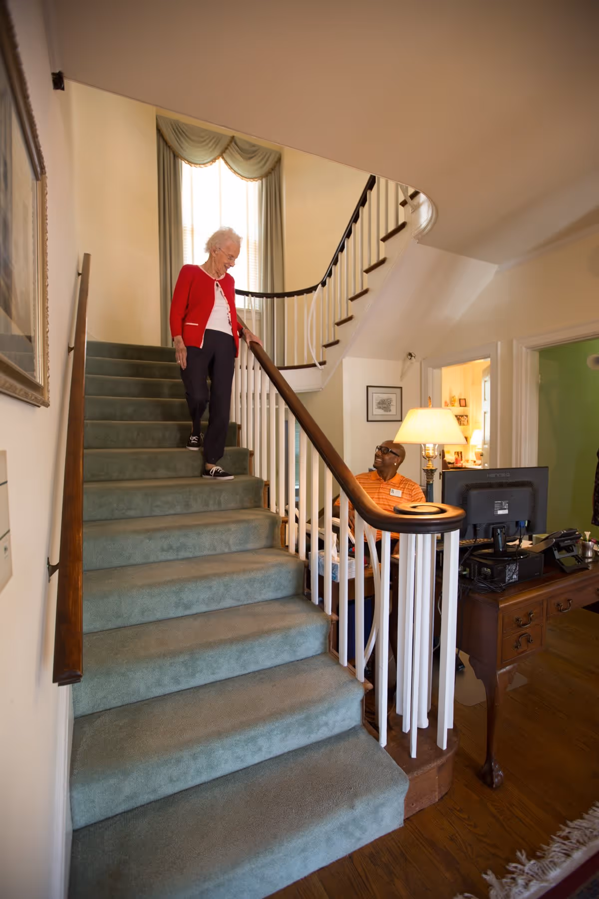 An elderly woman in a red cardigan and black pants is walking down a carpeted staircase with a wooden handrail inside a well-lit home or facility. A man wearing glasses and an orange striped shirt is sitting at a desk with a computer and lamp, looking up at her and smiling. The interior features light-colored walls, a large window with draped curtains, framed artwork, and wooden flooring.