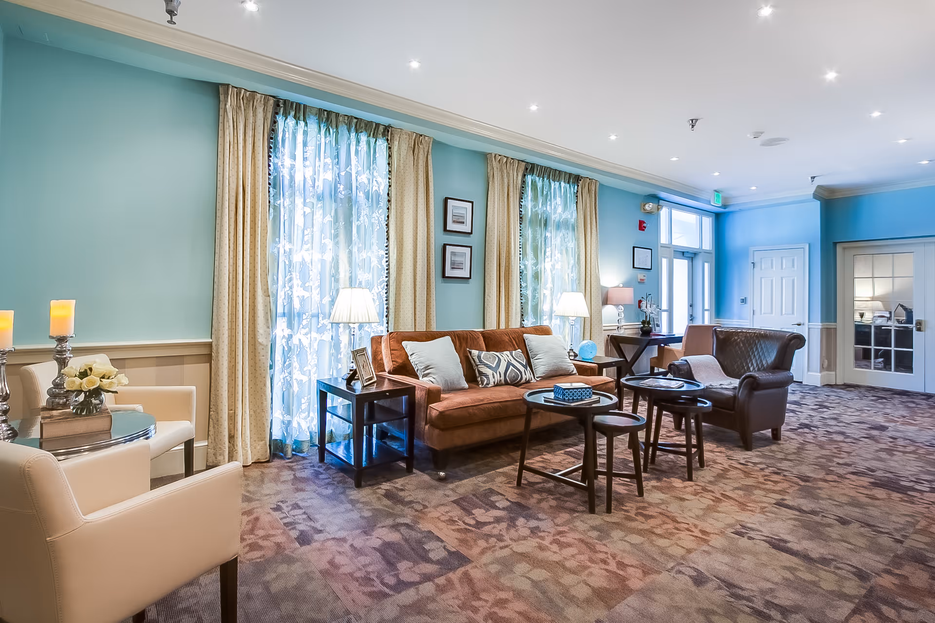 A bright and spacious senior living facility common area with teal walls and beige curtains over large windows. The room features a brown sofa with decorative pillows, a dark leather armchair, several small round tables, and cream-colored armchairs. There are table lamps, framed pictures on the walls, and a patterned carpet covering the floor.