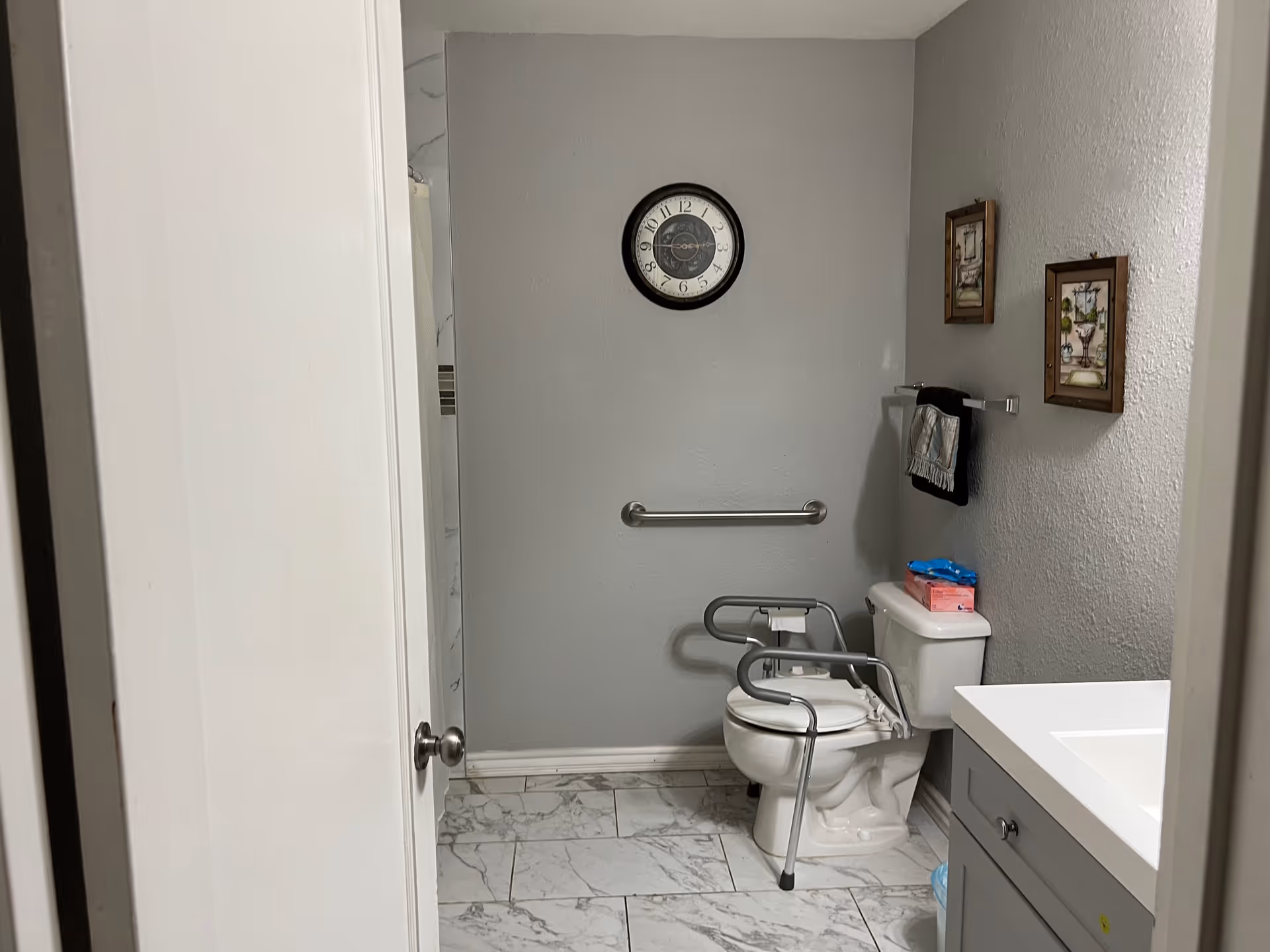 Accessible bathroom with a toilet, grab bars, sink, and a clock on the gray wall.