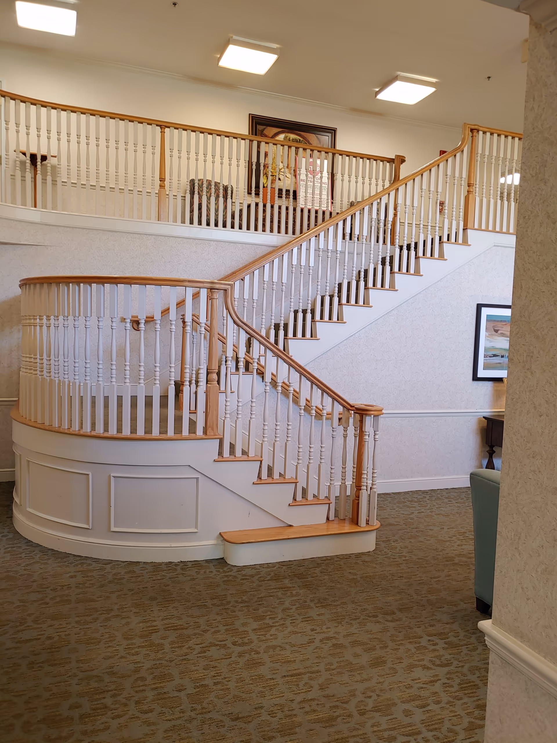 Interior view of a senior living facility showing a carpeted floor and a staircase with white spindles and wooden handrails leading to an upper level. The walls are light-colored with framed artwork and a small table visible in the background.