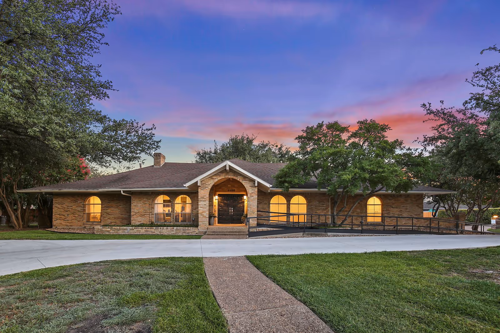 Exterior front view of a single-story brick assisted living building with arched windows, a ramped entrance, and illuminated interior at dusk.