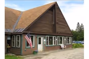 Exterior view of a brown wooden building with a steep triangular roof, several windows, a white door, an American flag mounted near the entrance, and a white chair placed outside. There are trees and parked cars visible in the background.