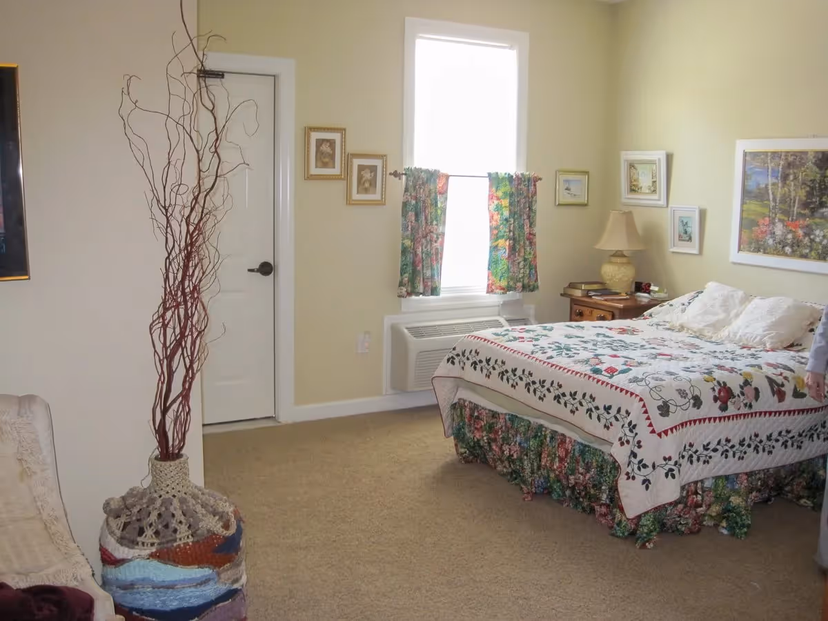 A cozy bedroom with a bed covered in a floral quilt and matching bed skirt. There is a window with floral curtains, a nightstand with a lamp and books, and several framed pictures on the walls. A decorative vase with tall branches is placed near a closed door.