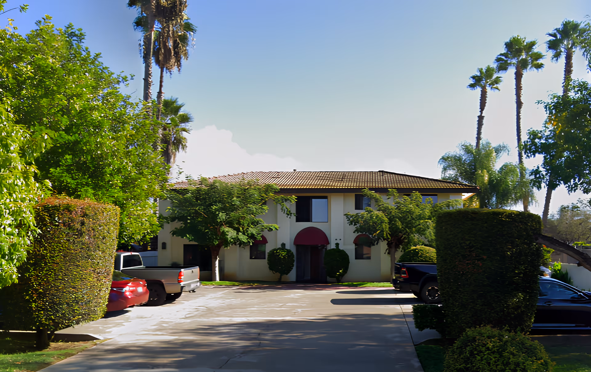 Front exterior view of a two-story building with a tiled roof and red awnings over the entrance and windows. The building is surrounded by neatly trimmed bushes and trees, with several parked cars along the driveway leading up to the entrance. Tall palm trees are visible in the background under a clear blue sky.