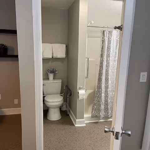 View into a small bathroom with a toilet, folded towels, a potted plant, and a shower with a patterned curtain.