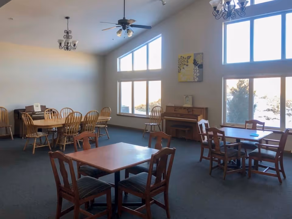 A spacious room with multiple wooden tables and chairs arranged for seating. Large windows let in natural light, and there are two pianos against the walls. The room has ceiling fans and chandeliers, with a carpeted floor and light-colored walls.