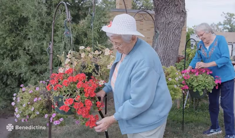 Two elderly women tending to hanging flower baskets outdoors in a garden area with green trees and grass. One woman is wearing a white hat and light blue jacket, while the other is dressed in a blue top and dark pants.