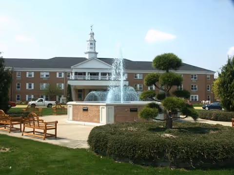 Exterior view of a large brick senior living facility building with a white cupola on the roof. In front of the building is a circular fountain with water spraying upwards, surrounded by a landscaped garden with trimmed bushes and uniquely shaped trees. Several wooden benches are placed on the paved walkway around the fountain.