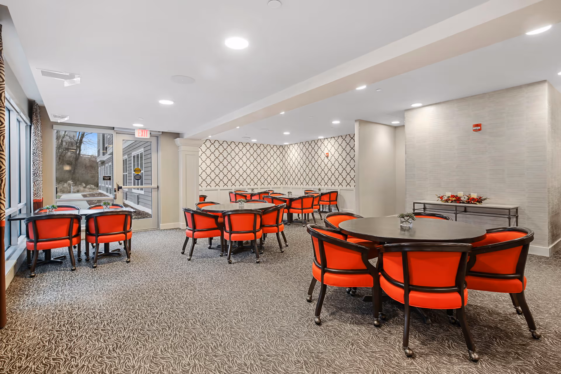 A bright and spacious dining room with multiple round tables surrounded by red cushioned chairs. The room features patterned carpet, a decorative wall with a geometric design, and large windows letting in natural light. There is a glass door leading outside and a console table with floral decorations against one wall.
