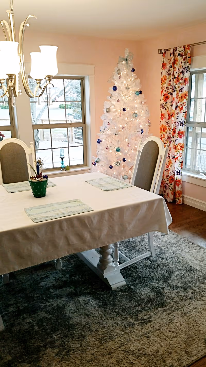 Dining room with a covered rectangular table and chairs, a decorated white Christmas tree by the windows, floral curtains and a chandelier.