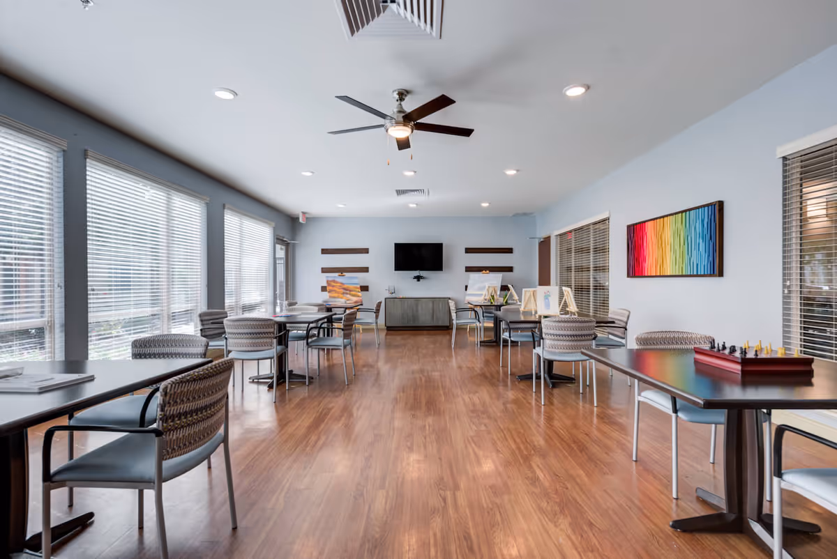A spacious, well-lit activity or common room with multiple tables and chairs arranged neatly. The room has large windows with blinds on the left side, a ceiling fan with lights, and a wooden floor. On the right wall, there is a colorful abstract painting, and on the far wall, a flat-screen TV is mounted above a cabinet. Some tables have art supplies and a chess set on them.