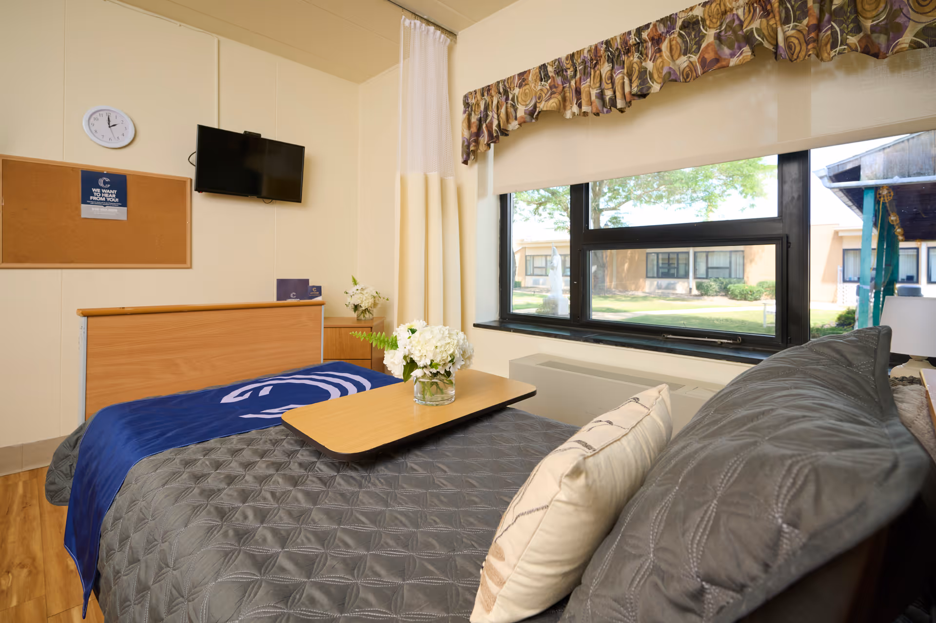 A cozy room in a rehabilitation and healthcare center featuring a hospital-style bed with a blue blanket and a gray quilted cover. A wooden overbed table holds a vase with white flowers. The room has a large window with a floral valance and a view of the outdoor courtyard. A wall-mounted TV, a clock, and a cork bulletin board are visible on the cream-colored walls.
