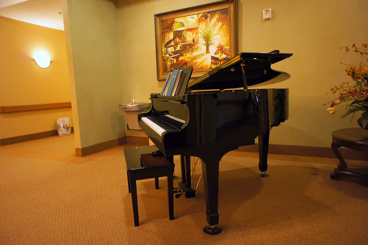 A black grand piano with a matching bench is placed in a warmly lit room with beige walls and carpet. Behind the piano, there is a framed painting depicting a piano and a person playing it. To the left, a water fountain is mounted on the wall, and to the right, a round table with a floral arrangement is visible.
