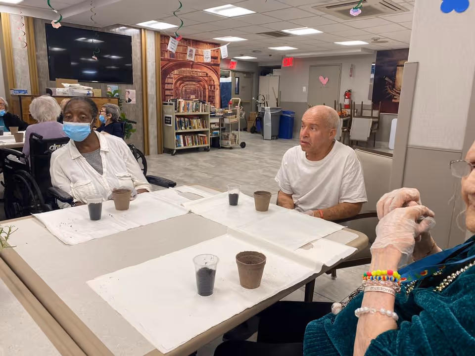 Several nursing home residents sit around a table with small cups and planting pots in a communal activity/dining room.