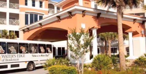 Exterior view of Water's Edge of Bradenton building entrance with a covered drop-off area, palm trees, and a shuttle bus parked nearby displaying the facility's name and slogan.