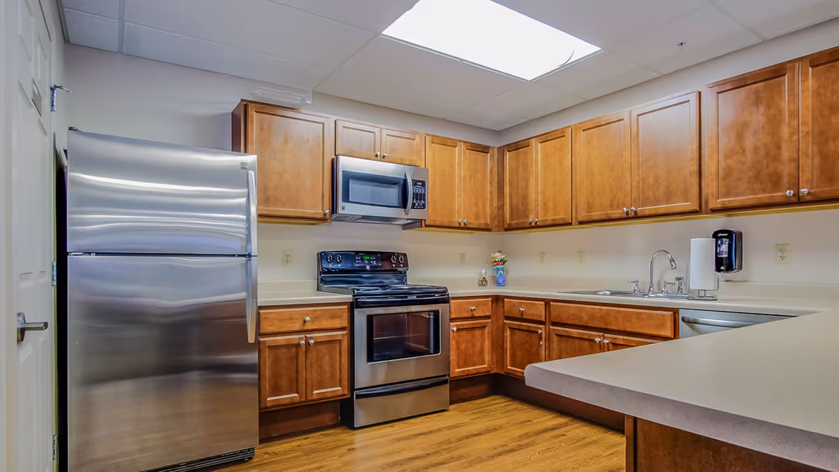 A clean and modern kitchen with wooden cabinets, stainless steel refrigerator, stove, microwave, and dishwasher. The kitchen has a light-colored countertop and a double sink with a faucet. There is a paper towel holder and a soap dispenser mounted on the wall. The floor is wooden and the ceiling has a large fluorescent light panel.