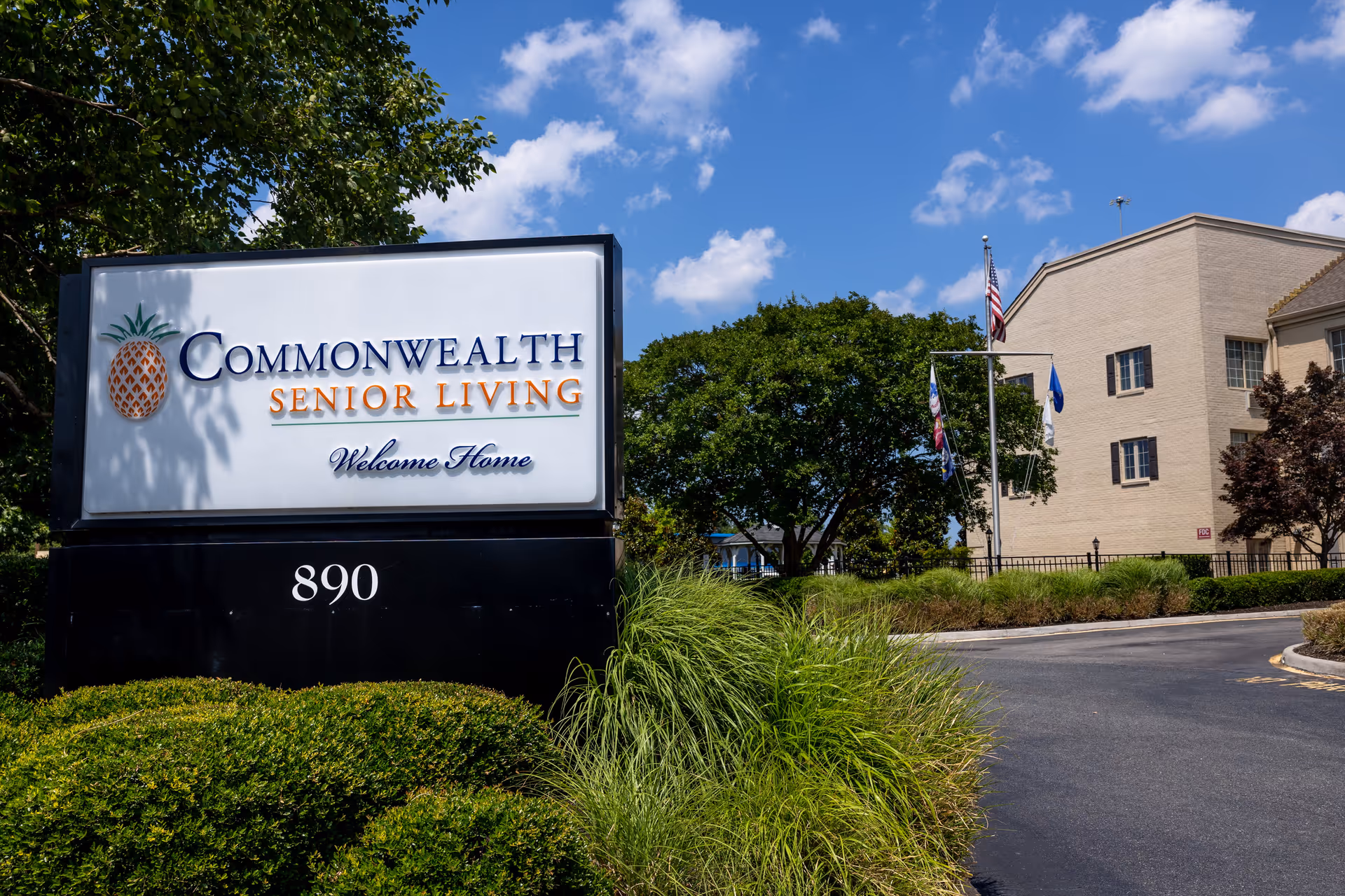 Outdoor view of the entrance sign for Commonwealth Senior Living at Leigh Hall with a large white sign featuring a pineapple logo and the text 'Commonwealth Senior Living Welcome Home' and the number 890. The background shows a beige building, trees, flags, and a clear blue sky with some clouds.