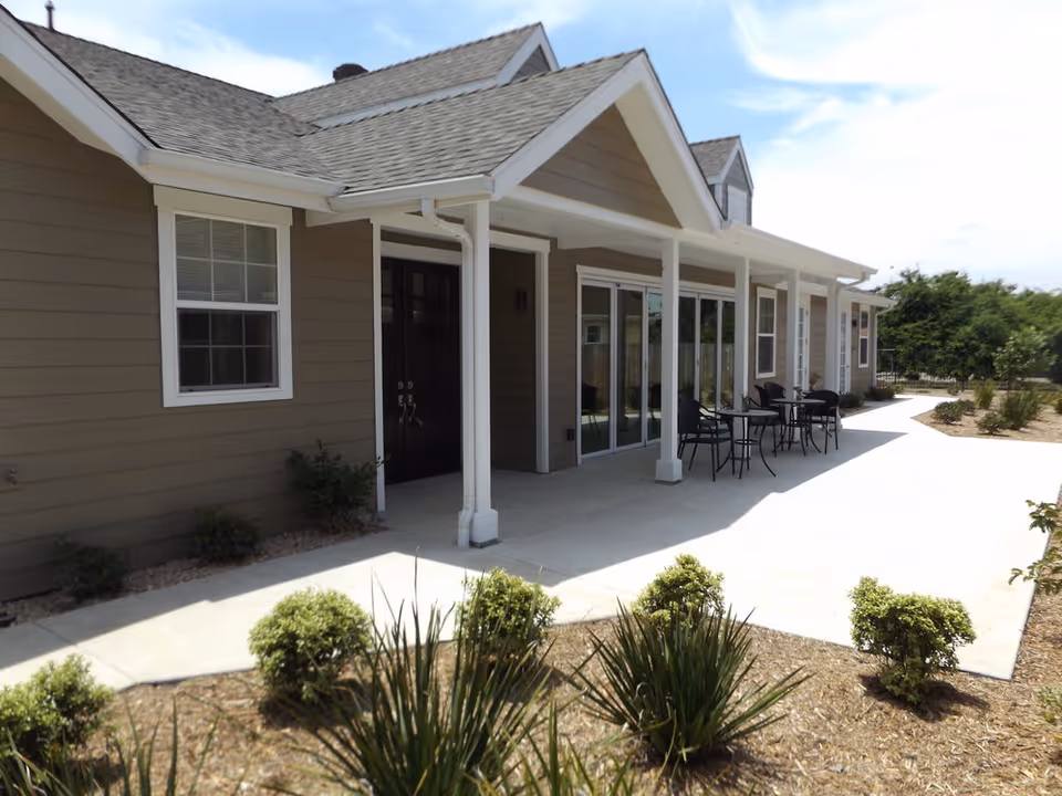 Exterior view of a single-story assisted living facility building with beige siding and a gray shingled roof. There is a covered patio area with white columns, outdoor tables and chairs, and a concrete walkway surrounded by landscaped bushes and plants under a partly cloudy sky.