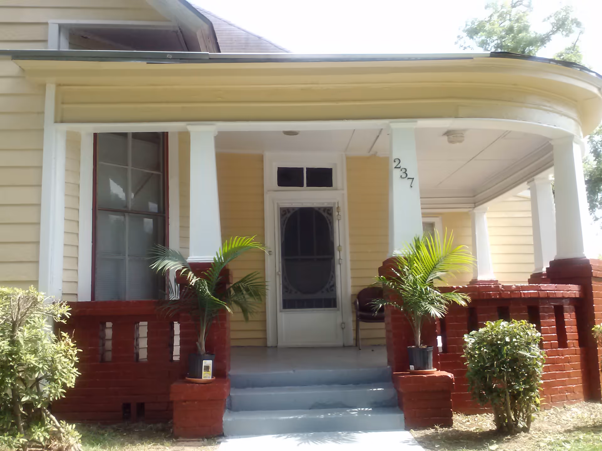 Front porch of a yellow house with white columns and red brick railings. Two potted plants are placed on either side of the steps leading to a white door with a decorative screen. The house number 237 is visible on one of the columns.