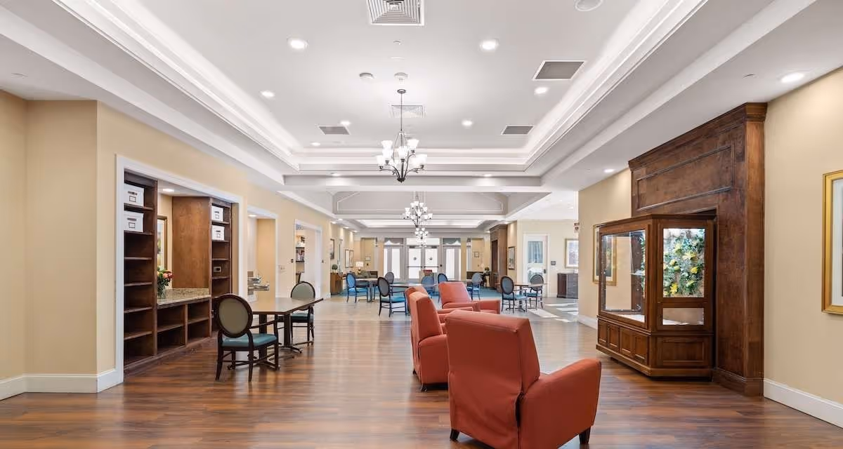 A spacious and well-lit interior common area in a senior living facility with wooden flooring, beige walls, and white tray ceilings with recessed lighting. The room features several red armchairs arranged in a row, multiple tables with chairs, built-in wooden shelves, and a large wooden display cabinet with glass panels. The area appears clean and inviting with natural light coming through windows in the background.