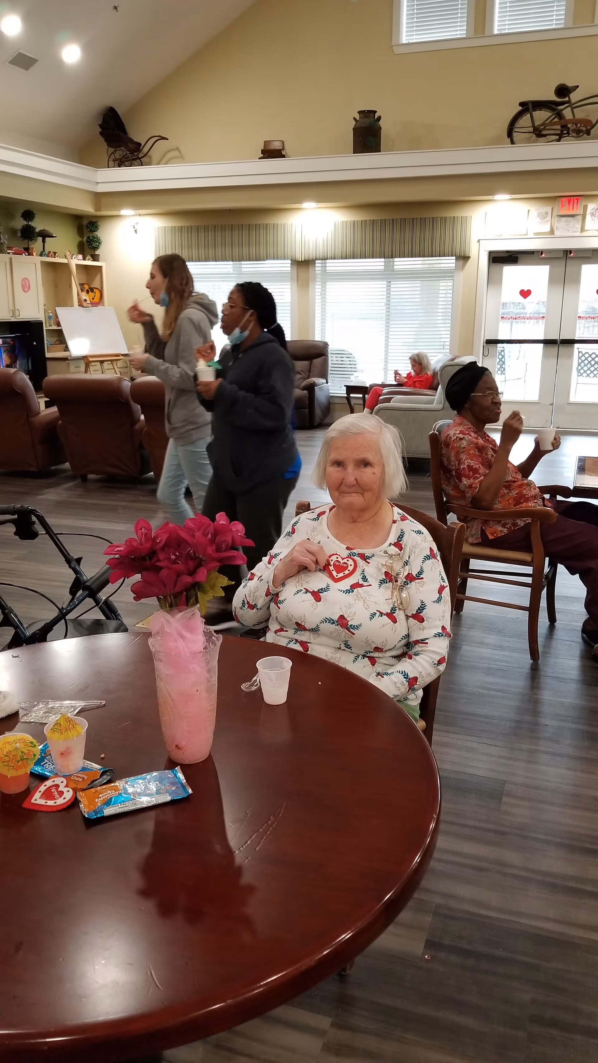 An elderly woman with white hair wearing a floral shirt sits at a round wooden table with a pink vase of flowers and snacks in a spacious common area. Behind her, two women walk by holding cups, and two other elderly women sit in chairs near large windows and a glass door. The room has high ceilings and comfortable seating.