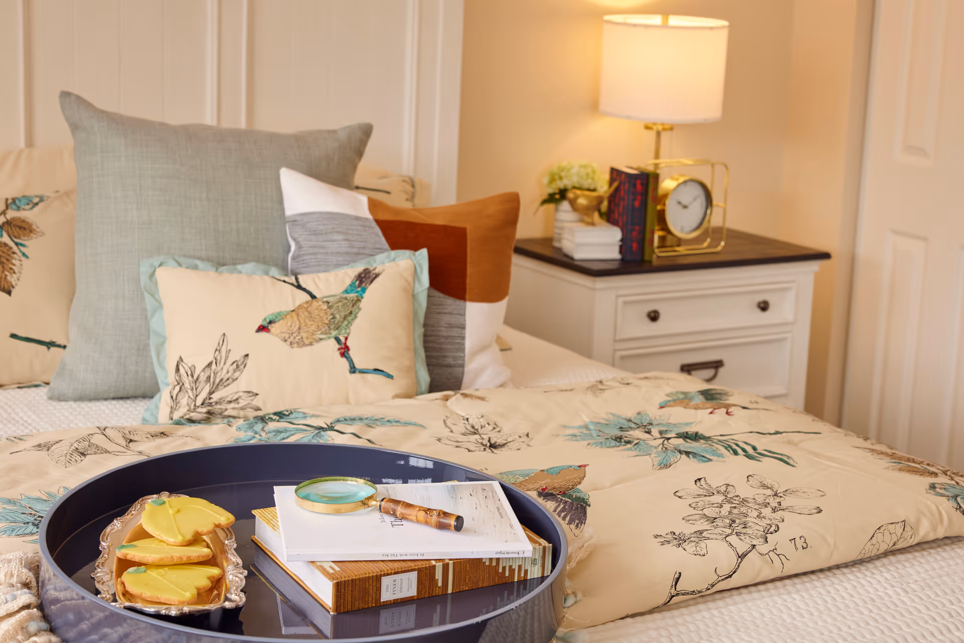 A cozy bedroom with bird-patterned bedding and pillows, a bedside table with a lamp and clock, and a tray on the bed holding cookies and books.