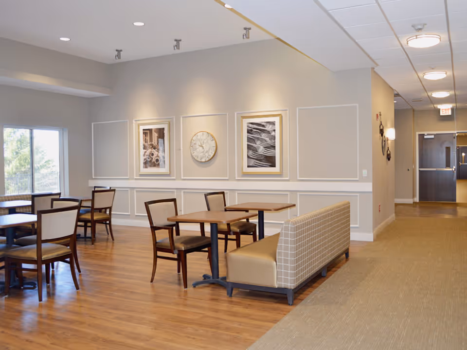 A bright and clean dining area in a senior living facility with wooden floors, several tables and chairs, a cushioned bench, framed artwork, and a large window letting in natural light.