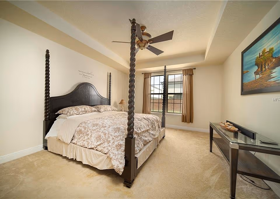 A spacious bedroom with a large four-poster bed featuring dark wood posts and a floral patterned bedspread. The room has beige carpet, cream-colored walls, a ceiling fan with wooden blades, a window with brown curtains, and a glass-top console table with decorative items and a painting on the wall above it.