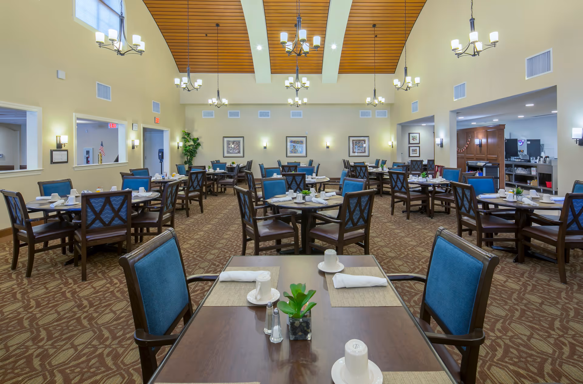 A spacious dining room with multiple round tables and blue cushioned chairs arranged neatly. Each table is set with white cups, saucers, napkins, and small plants as centerpieces. The room features a high ceiling with wooden paneling and several hanging chandeliers providing warm lighting. The walls are light-colored with framed artwork and wall sconces. There is an open kitchen area visible on the right side of the room.