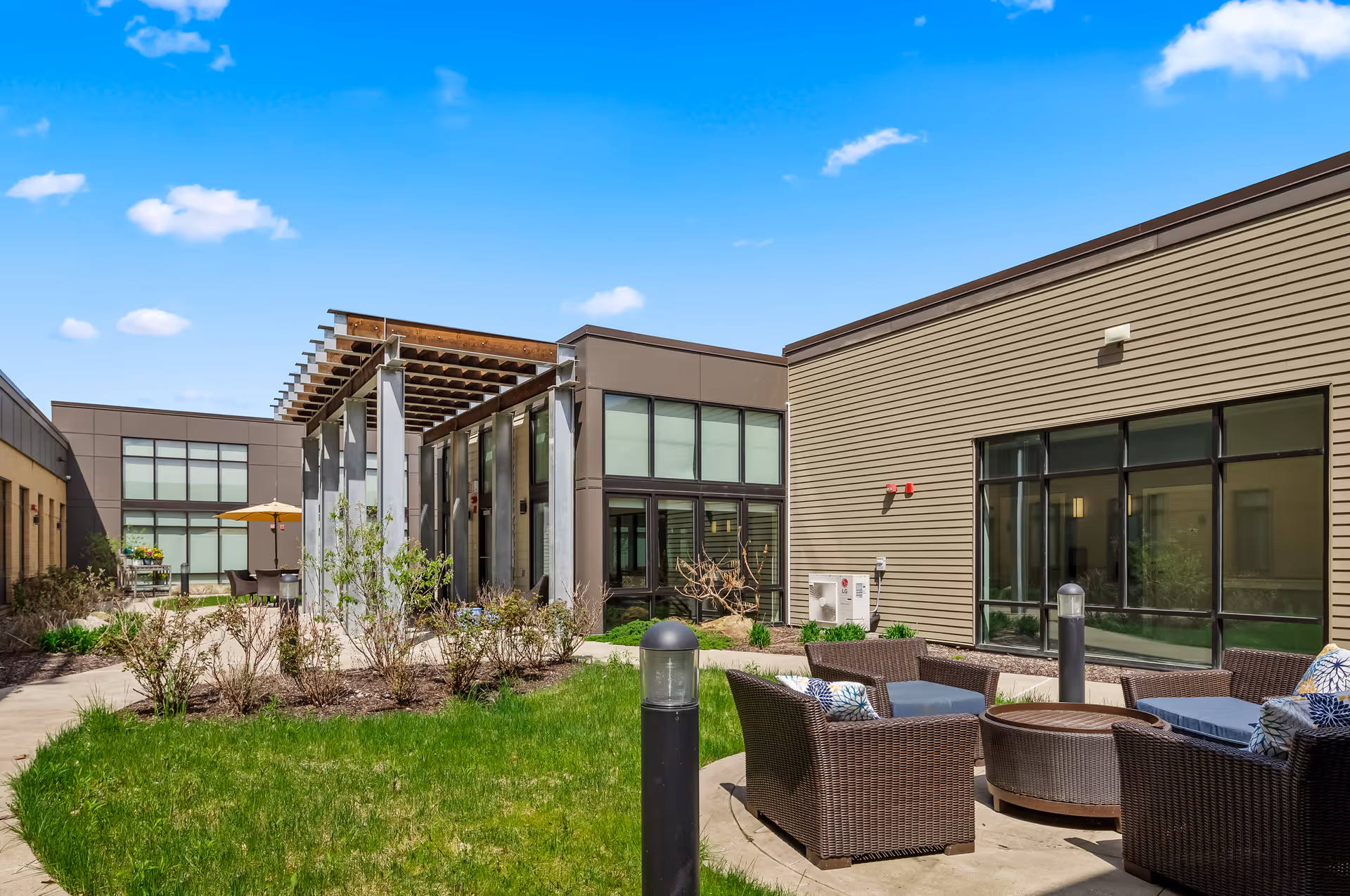 Outdoor courtyard area at Ridgecrest of McCandless featuring modern building exteriors with large windows, a seating area with wicker chairs and cushions around a fire pit, landscaped greenery, and a pergola structure under a bright blue sky with scattered clouds.