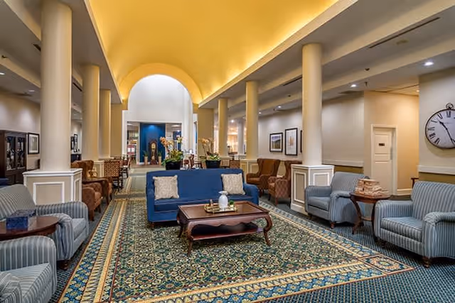 A spacious and elegant common area with a high arched ceiling and soft lighting. The room features a large patterned carpet, multiple upholstered armchairs and sofas arranged around wooden coffee tables, and decorative columns along the sides. The walls are adorned with framed artwork and a large clock is visible on the right side. The area appears welcoming and comfortable, suitable for socializing or relaxing.