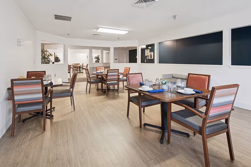 A bright dining room in a senior living facility with several wooden tables and chairs arranged neatly. Each table is set with plates, glasses, and blue napkins. The room has light-colored wooden flooring, white walls, and large rectangular windows or openings along one wall. In the background, another dining area is visible through the openings.