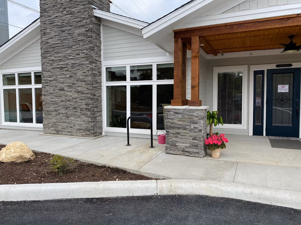 Exterior view of the entrance to BeeHive Homes of Crownridge Assisted Living, featuring a stone pillar, large windows, a blue door with a glass panel, a covered porch with wooden beams, and potted plants near the entrance.