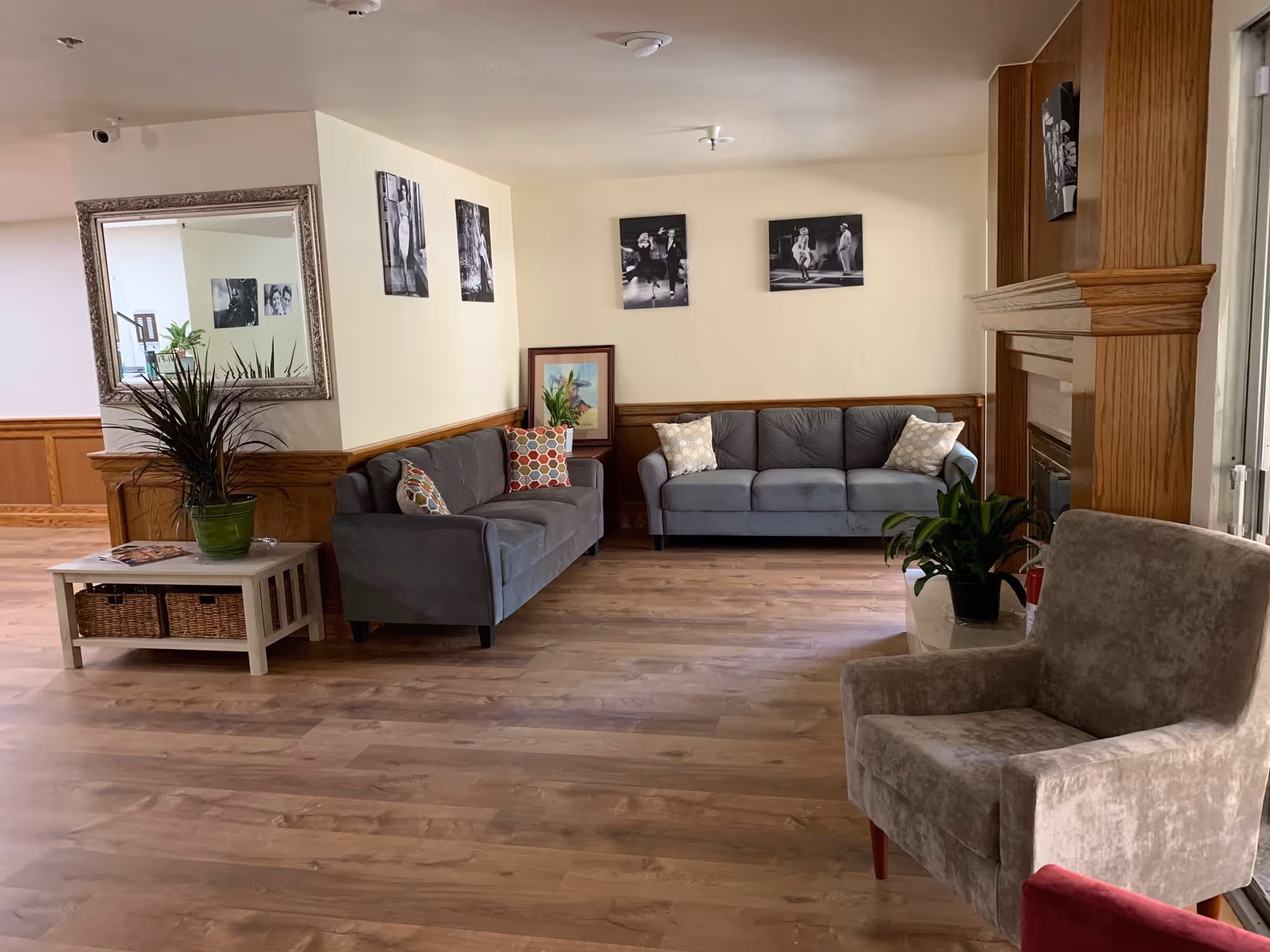 A cozy living room area with two gray sofas adorned with patterned pillows, a beige armchair, and a small white table with a green potted plant. The room features wooden flooring, wood paneling on the lower walls, a large mirror, black and white photos on the walls, and a fireplace with a wooden mantel.