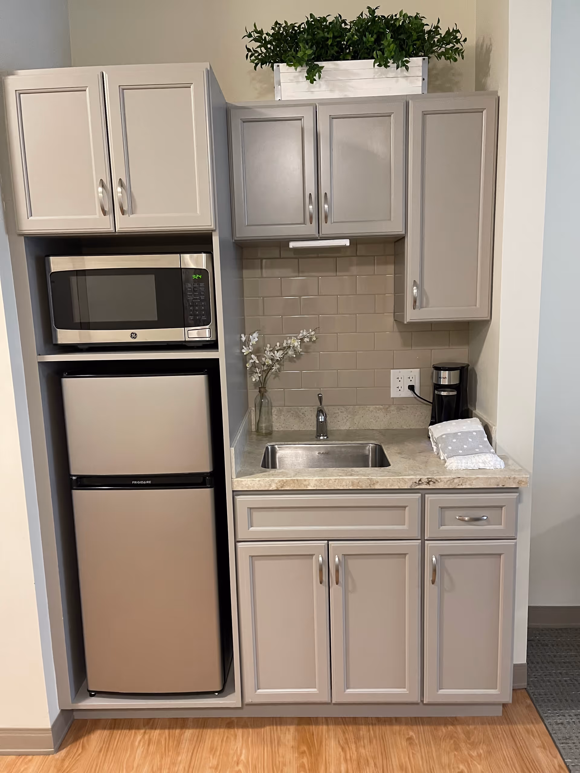 A small kitchenette area with light gray cabinets, a stainless steel microwave, a compact refrigerator, a sink with a faucet, a coffee maker, and a small vase with white flowers. There is a green plant in a white rectangular planter on top of the cabinets.