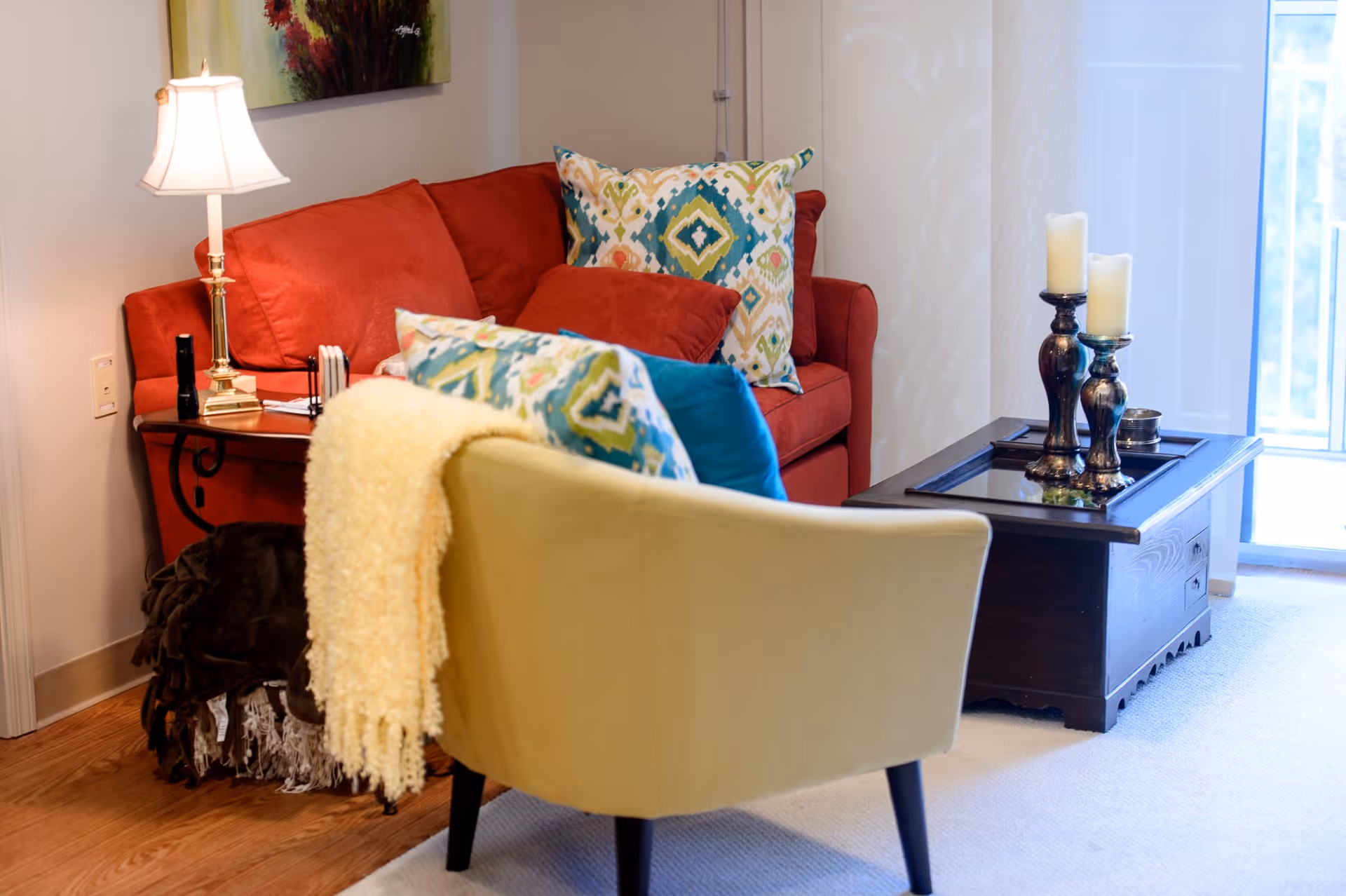 Cozy living room with a red sofa, patterned throw pillows, a yellow accent chair draped with a blanket, a side table lamp, and a wooden coffee table with candles.