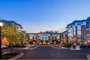 Exterior view of a multi-story senior living facility at dusk with well-lit pathways, landscaped trees, and a circular driveway in front of the building.
