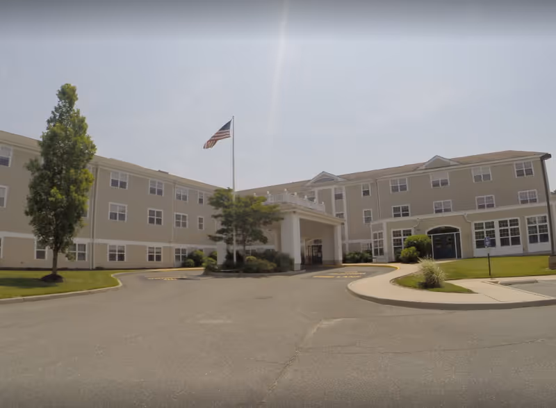 Exterior view of a three-story assisted living facility building with beige siding and white trim. There is a covered entrance with columns, an American flag on a flagpole, and landscaped greenery including trees and bushes around the driveway and entrance area.