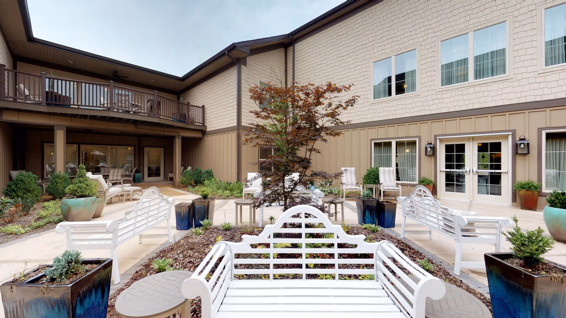 Outdoor courtyard area of a senior living facility with white benches, potted plants, a small tree in the center, and a two-story building with windows and doors surrounding the courtyard.
