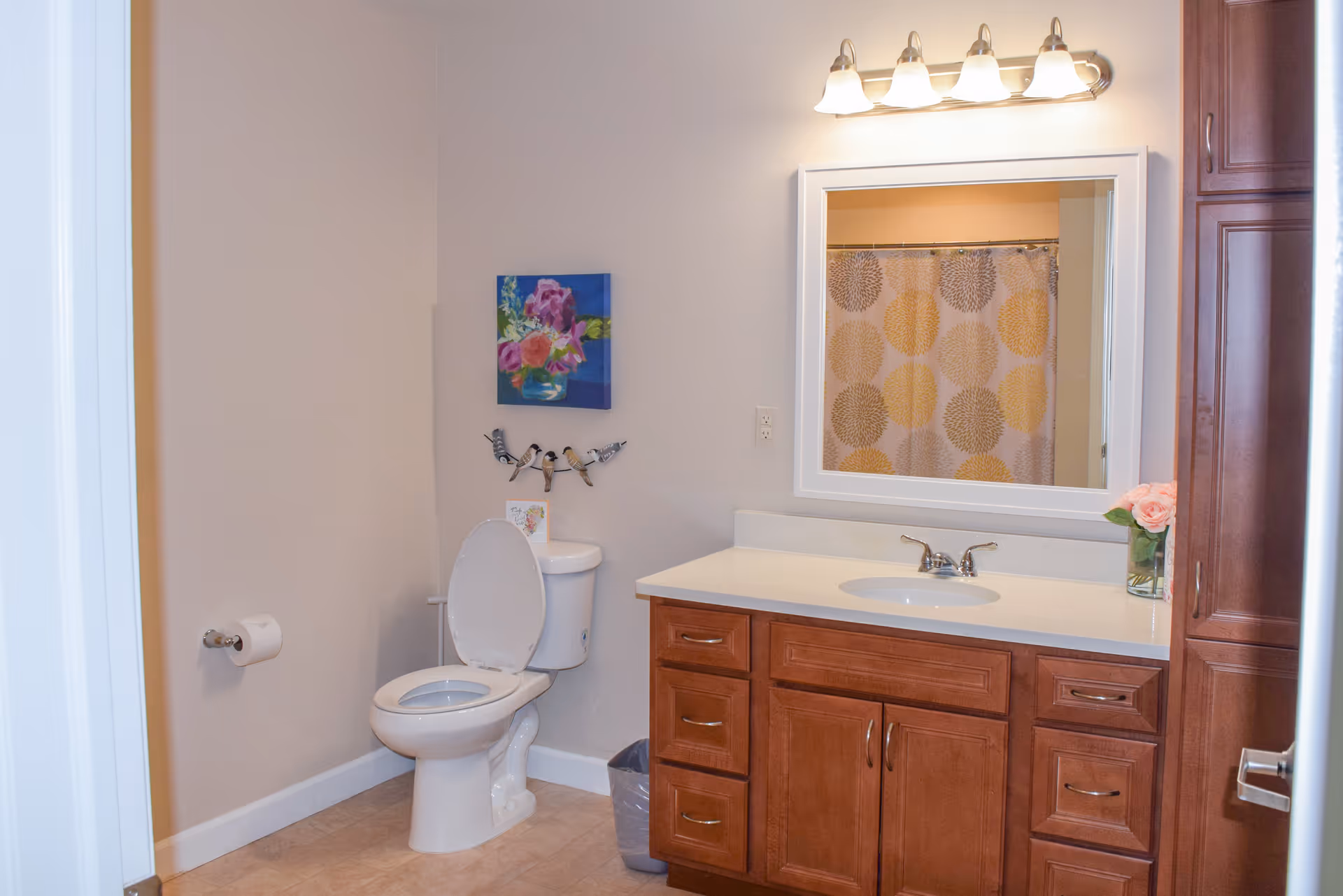 A clean bathroom with a white toilet with the lid open, a toilet paper holder on the wall, a wooden vanity with a white countertop and sink, a large mirror above the sink, a light fixture with four bulbs above the mirror, a shower curtain with circular patterns reflected in the mirror, a small trash bin, a floral painting on the wall, and a vase with pink flowers on the countertop.