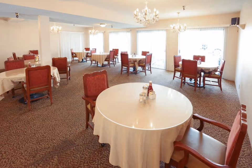 Bright dining room with round tables covered in white tablecloths, red upholstered chairs, chandeliers, and large windows.
