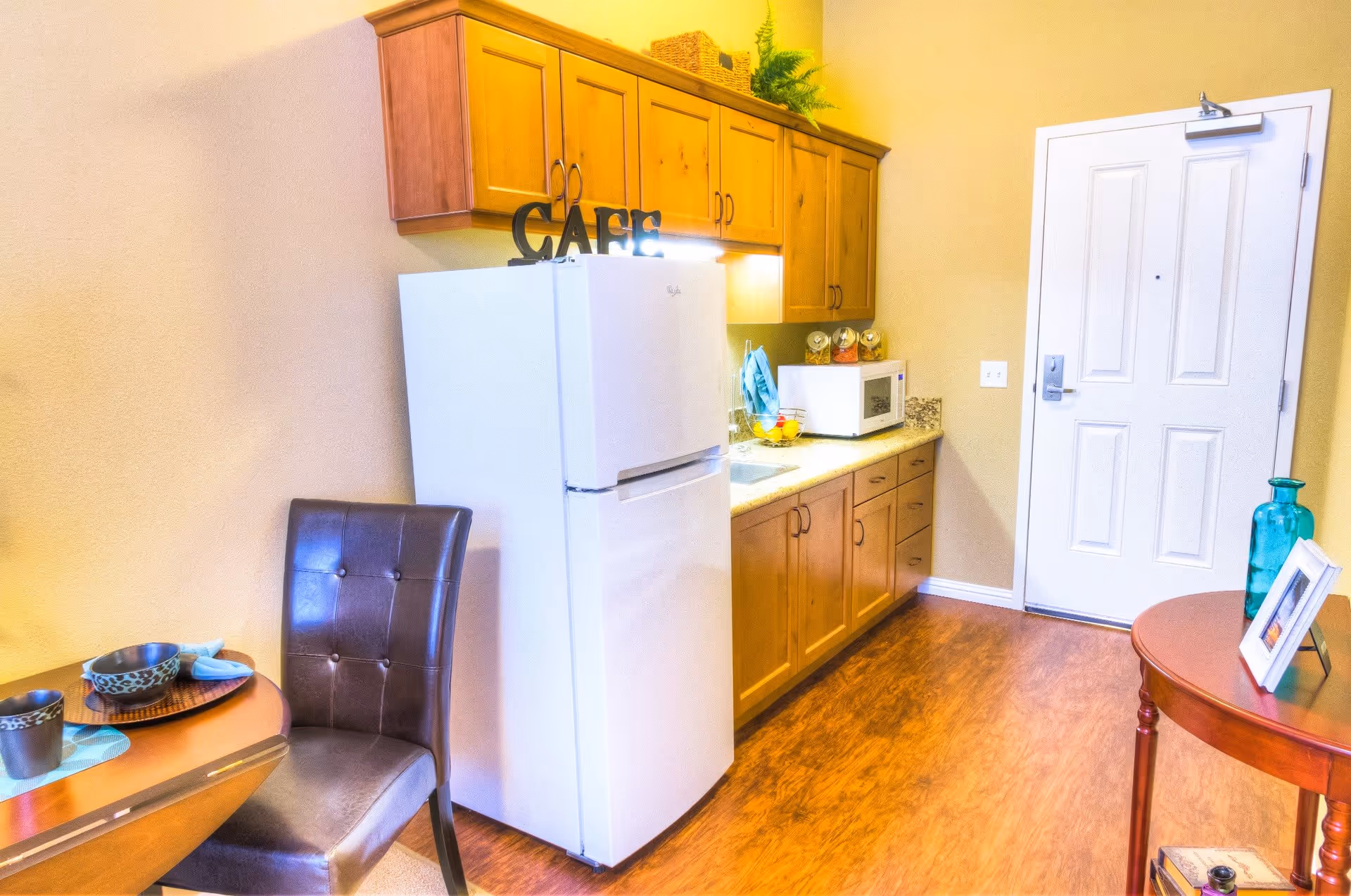 Interior view of a small kitchen area with wooden cabinets, a white refrigerator with a decorative 'CAFE' sign on top, a microwave on the countertop, and a round table with a brown leather chair. The floor is wooden, and there is a white door in the background. A small round side table with a blue vase and a framed photo is also visible.