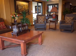 A cozy living room area in a senior living facility with two armchairs featuring patterned cushions, a wooden coffee table with decorative items, a side table with a lamp and flowers, and framed artwork on the walls.